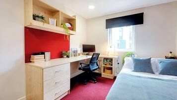 A bedroom with a bed, a desk, and a computer. There is a window with blinds, a shelf with plants, and a red accent wall.