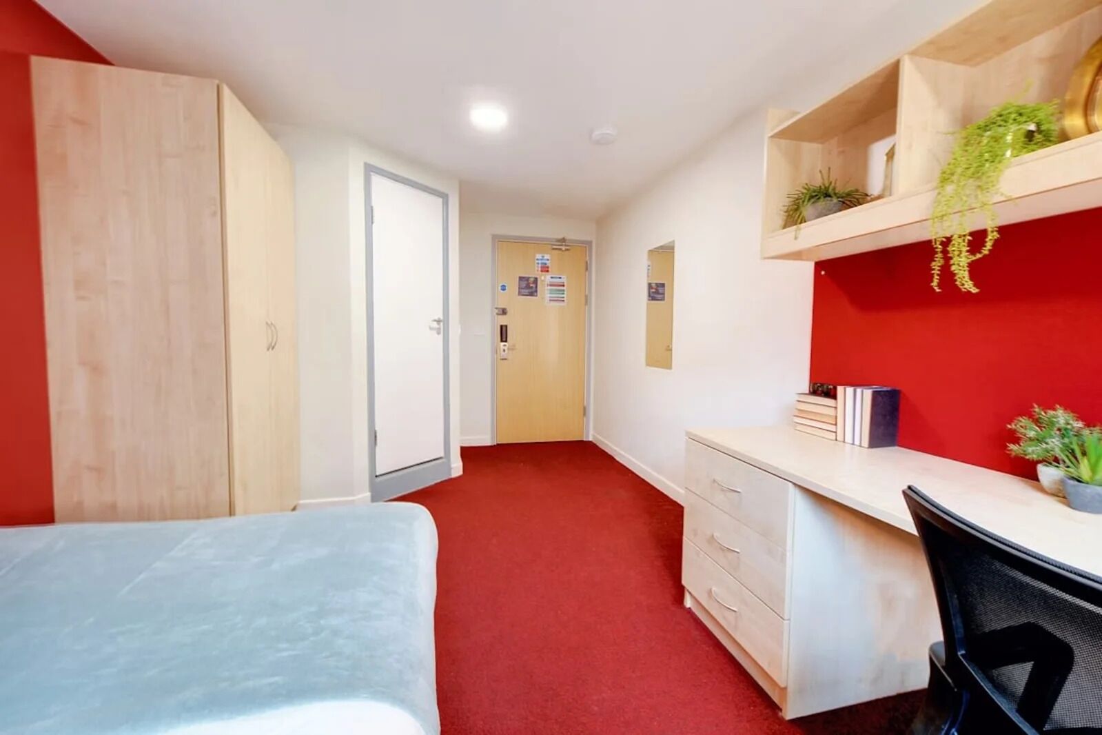Um quarto de estudante com uma cama, uma escrivaninha, uma cadeira, um guarda-roupa e um chão coberto por um tapete vermelho.