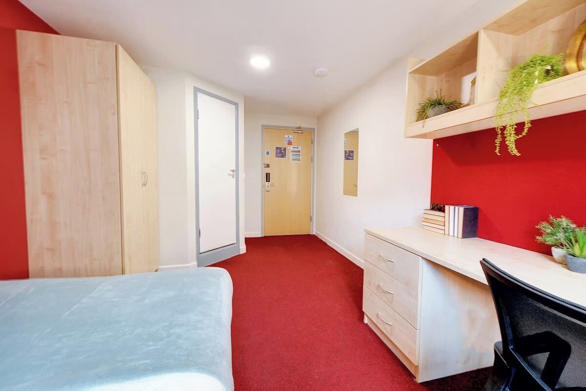 A room with a bed, a desk, a chair, a wardrobe, and a red carpet.