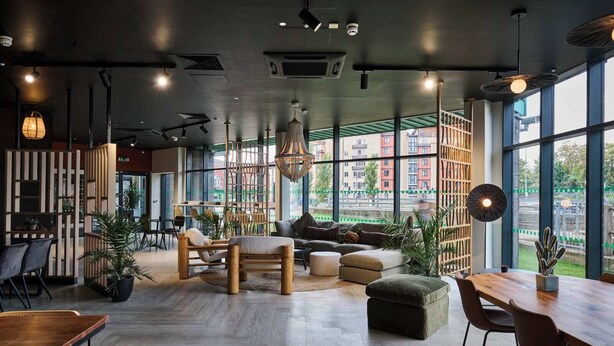 A modern interior with a wooden floor, a seating area with sofas and chairs, large windows offering a view of buildings, and hanging pendant lights. - For Students Only Nottingham Two (Nottingham)