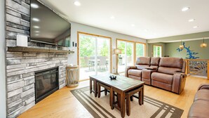 A living room with a stone fireplace, a flat-screen TV, a brown leather sofa, and a dining area with a table and chairs. - Deep Creek Escape: w/ Theater, Fire Pit & Dock (Swanton)
