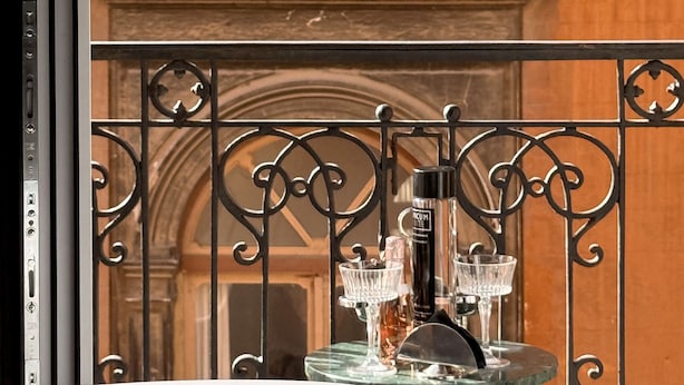 A balcony with a wrought iron railing, a glass table with a bottle and glasses, and a view of a building with arched windows. - Unicum Suite Grande Archivio Napoli  (Naples)
