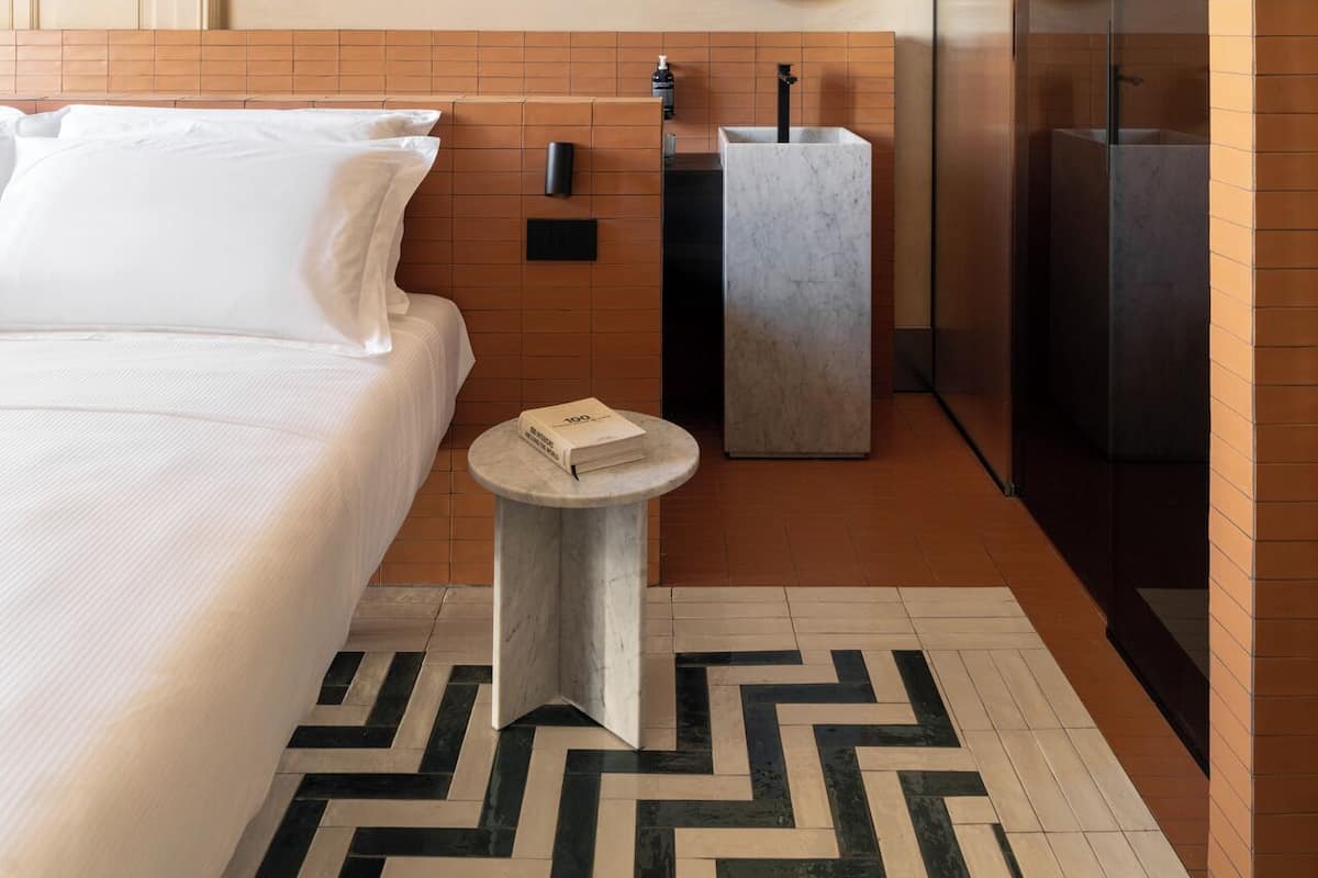 A modern hotel room with a large bed, a marble side table, a round mirror, and a shower area.