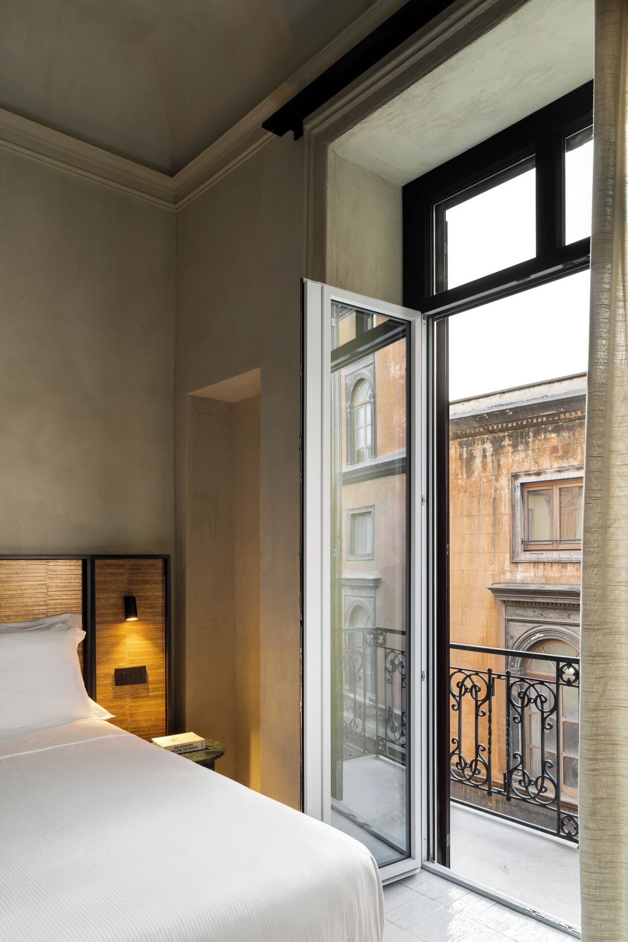 A hotel room with a bed, a nightstand, a window with a view of a building, and a balcony with a wrought iron railing.