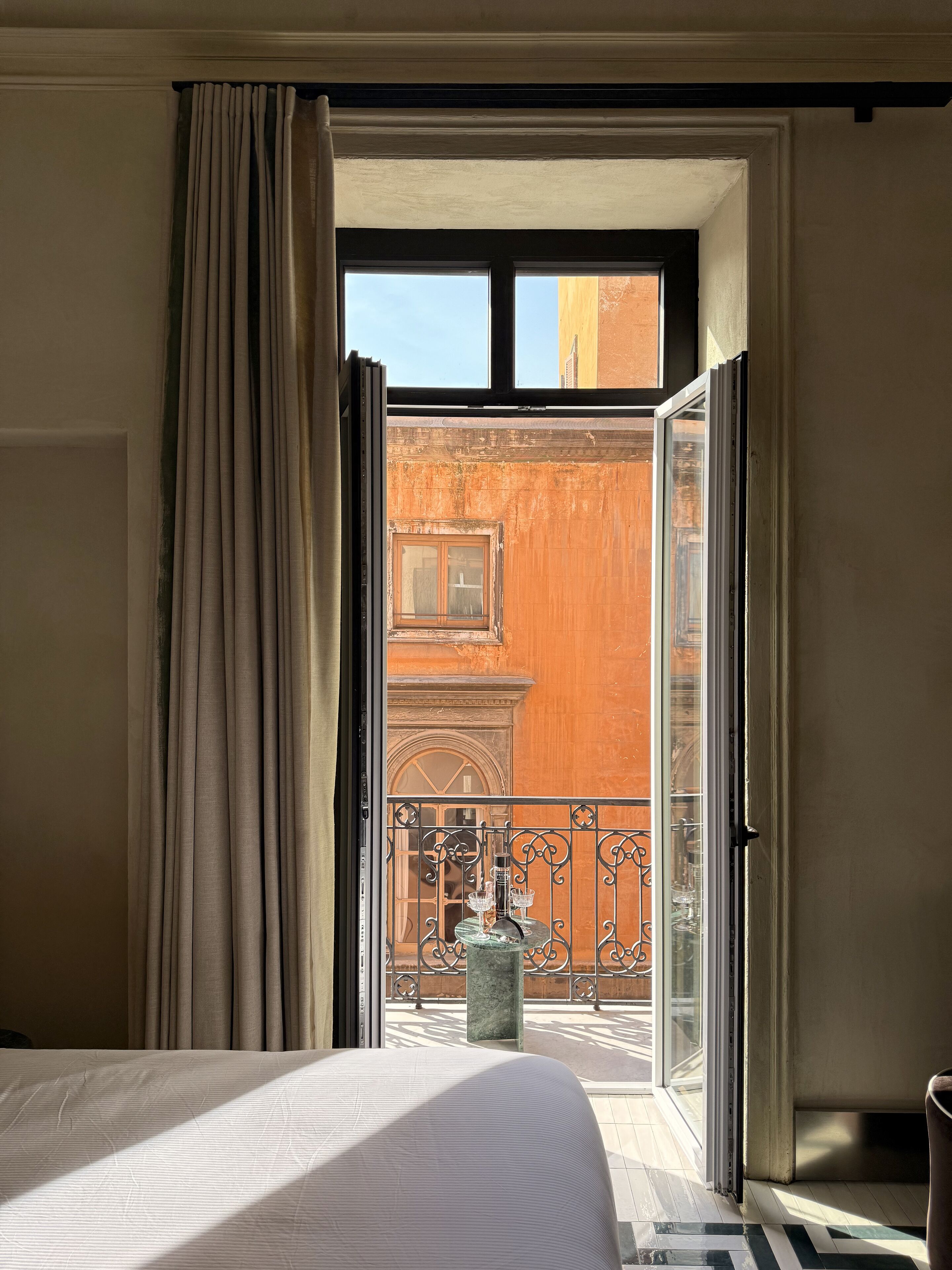 A hotel room with an open sliding door leading to a balcony with a view of a building and a metal railing.