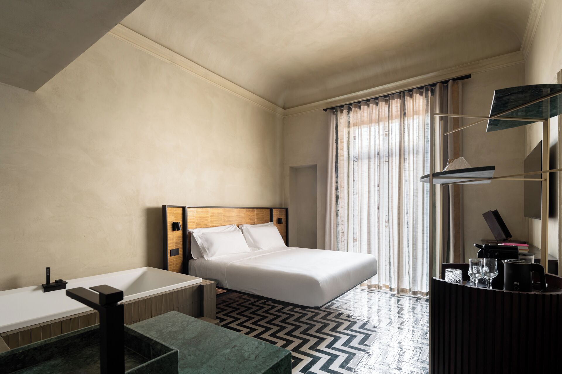A hotel room with a bed, a bathtub, a mirror, and a window with curtains.