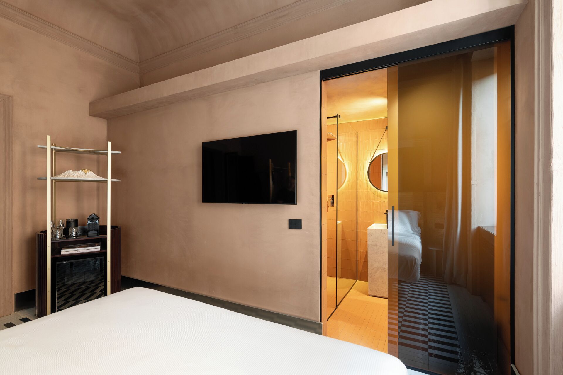A hotel room with a bed, a TV mounted on the wall, a wardrobe, and a bathroom visible through a glass door.