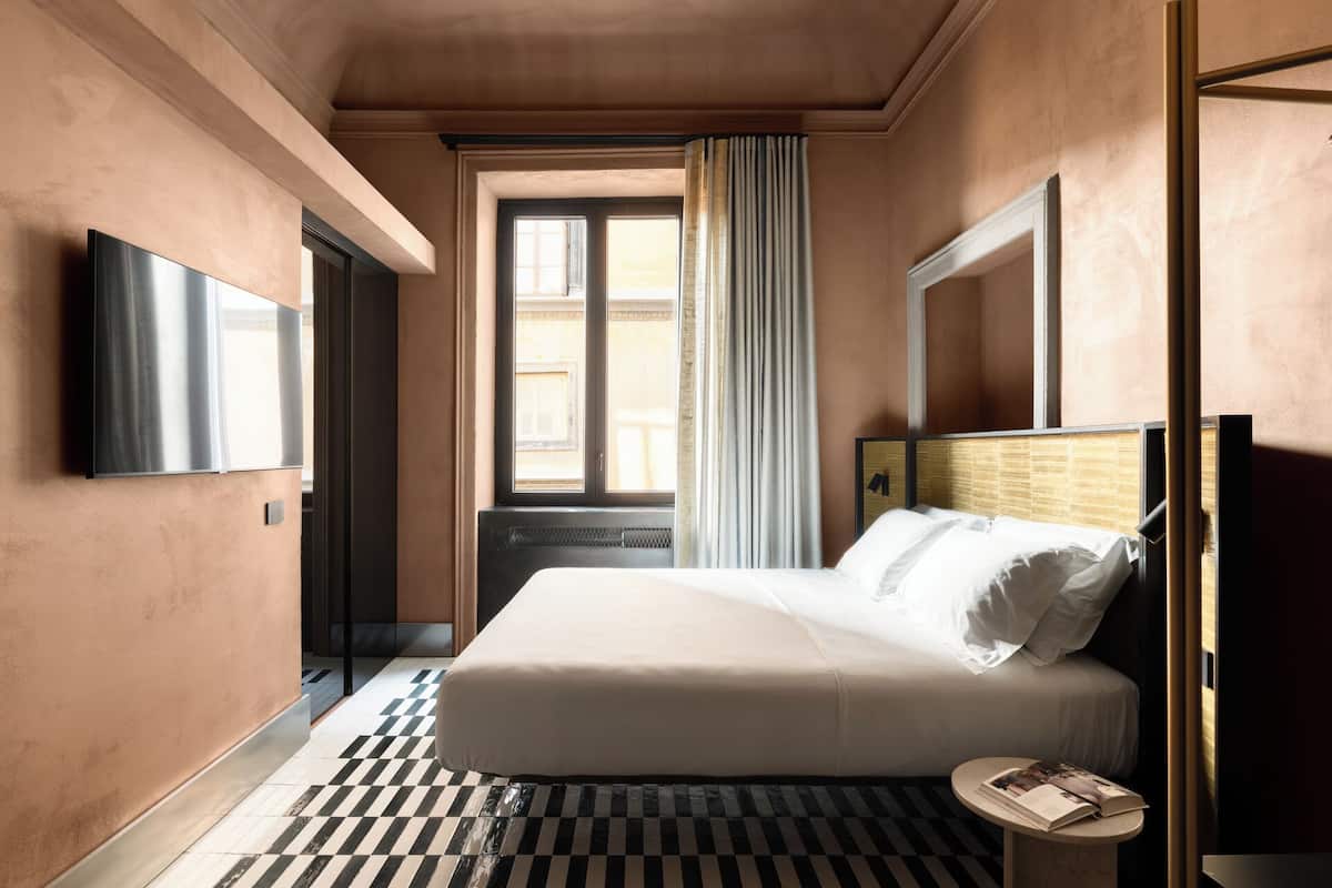 A modern hotel room with a large bed, a small bedside table, a mirror, and a window with curtains.