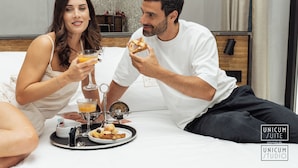 A couple sitting on a bed with breakfast items including orange juice, a plate of food, and a coffee maker. - Unicum Suite Grande Archivio Napoli  (Naples)