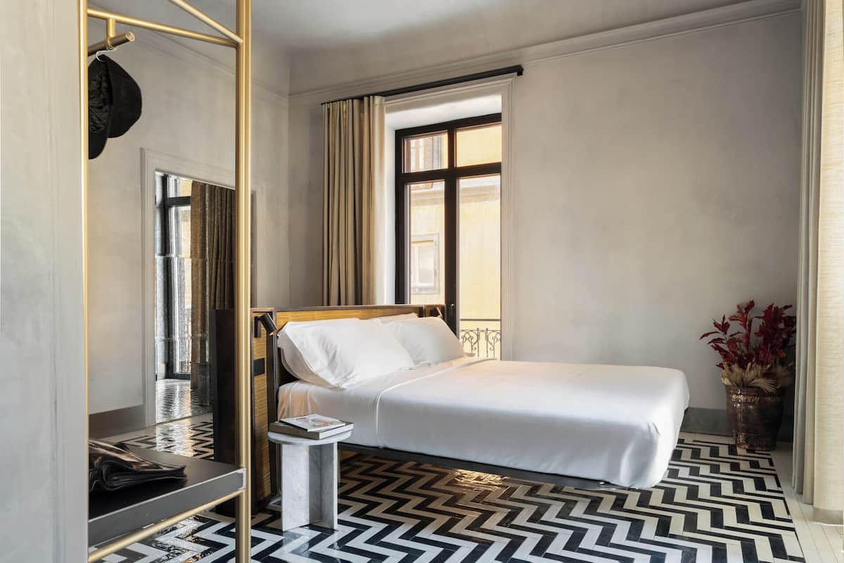 A hotel room with a bed, a bedside table, a mirror, a window with curtains, and a patterned floor.