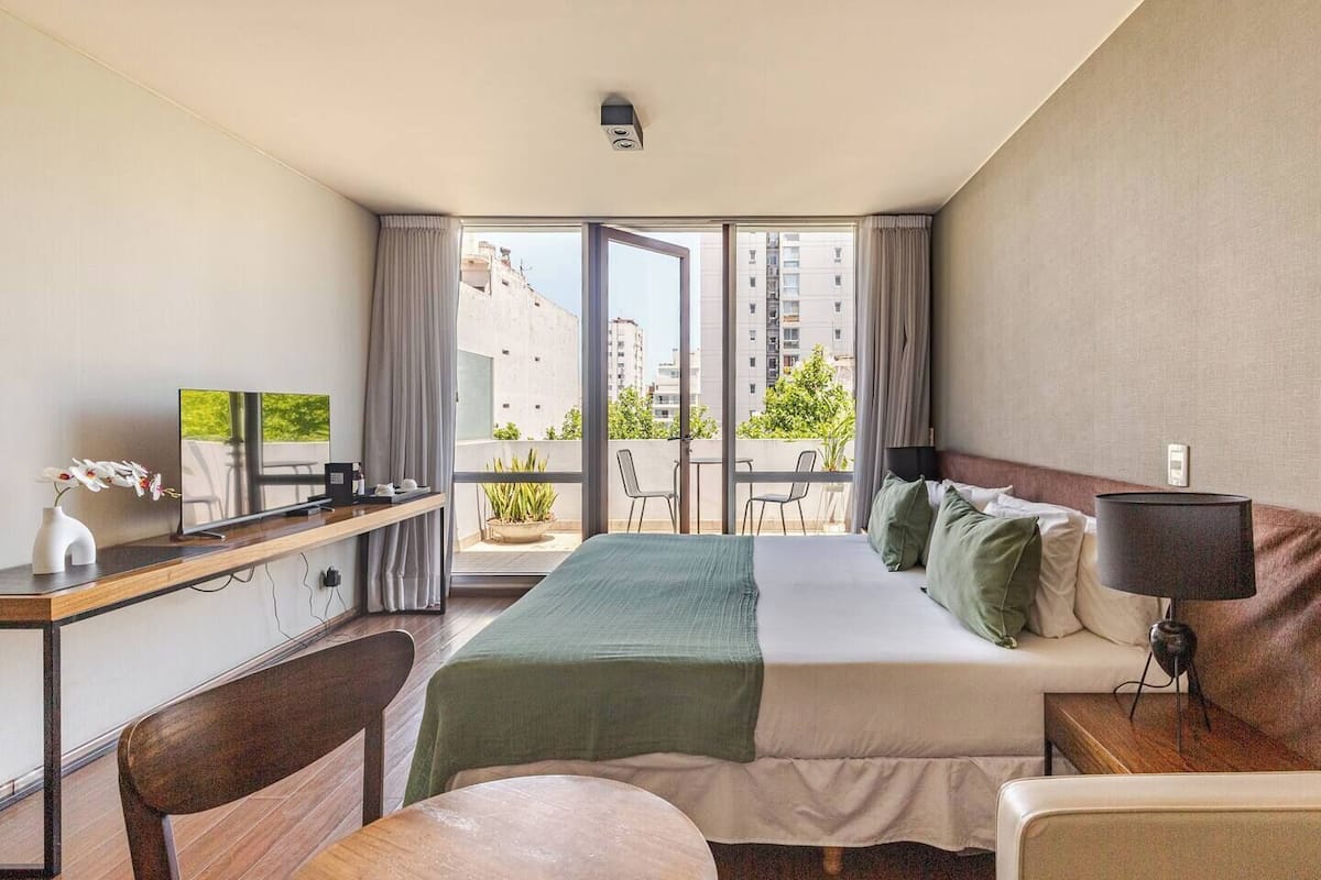 A hotel room with a bed, a desk with a TV, a balcony with a view of buildings and greenery.
