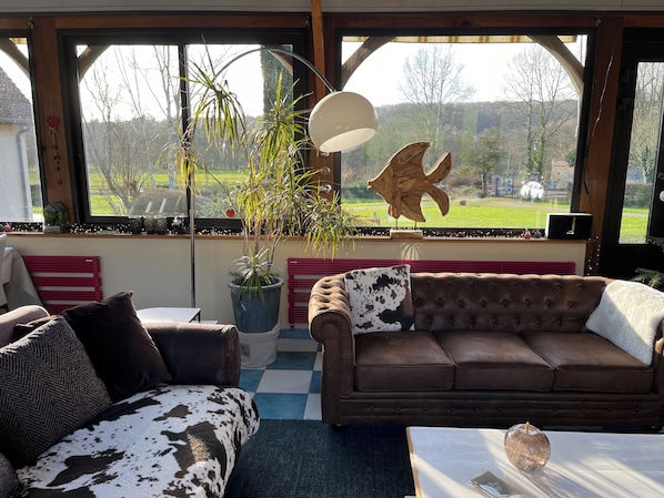 A cozy living room with a brown leather sofa, a patterned armchair, and a large window offering a view of a green landscape.
