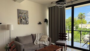 Ein modernes Wohnzimmer mit einer Couch, einem Holztisch, einem kleinen Beistelltisch, einer Stehlampe und einem großen Fenster mit Blick auf Grünflächen und Gebäude.