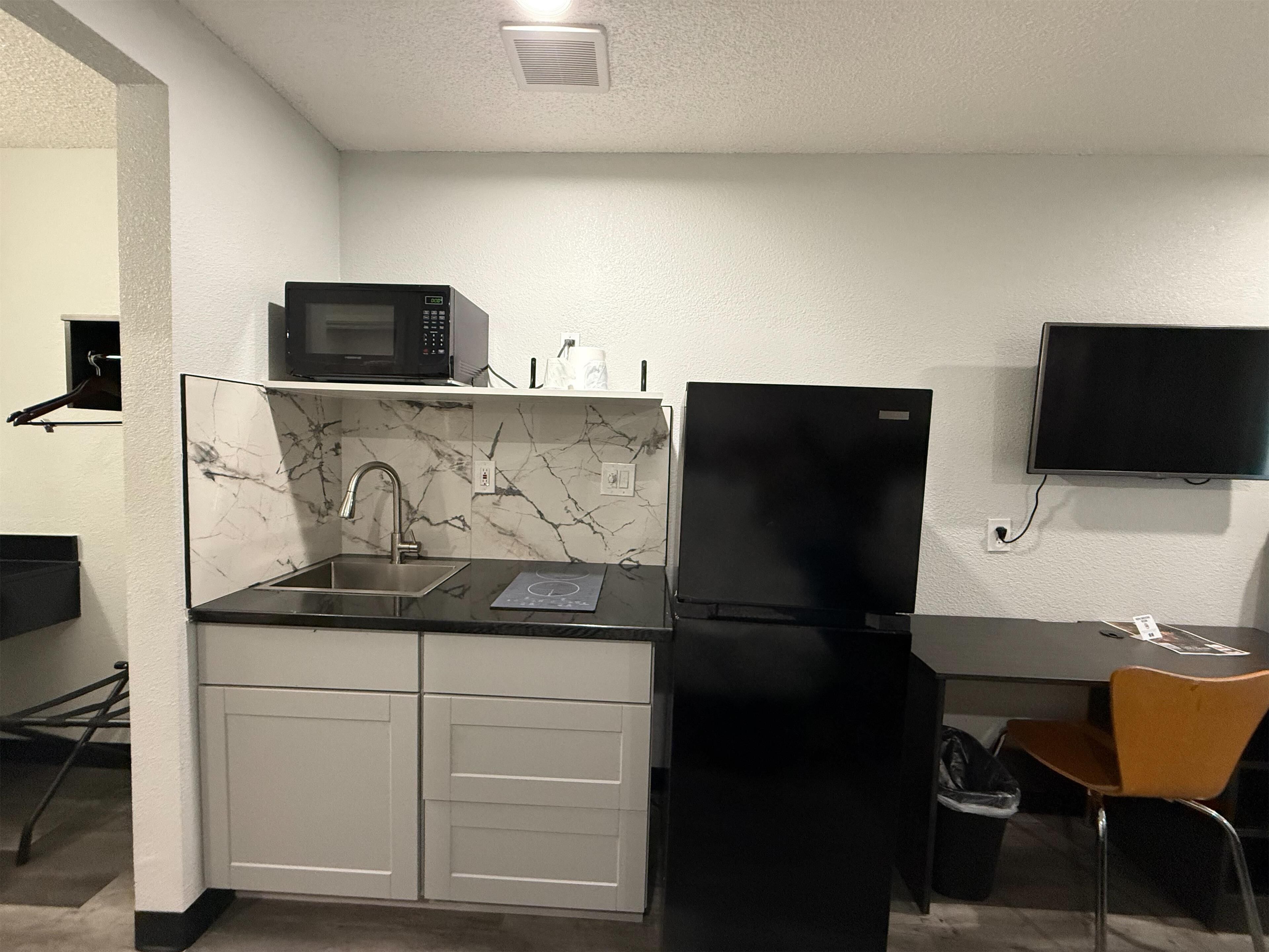 A compact kitchen with a black refrigerator, a microwave, and a sink. There is a small table with a chair and a TV mounted on the wall.