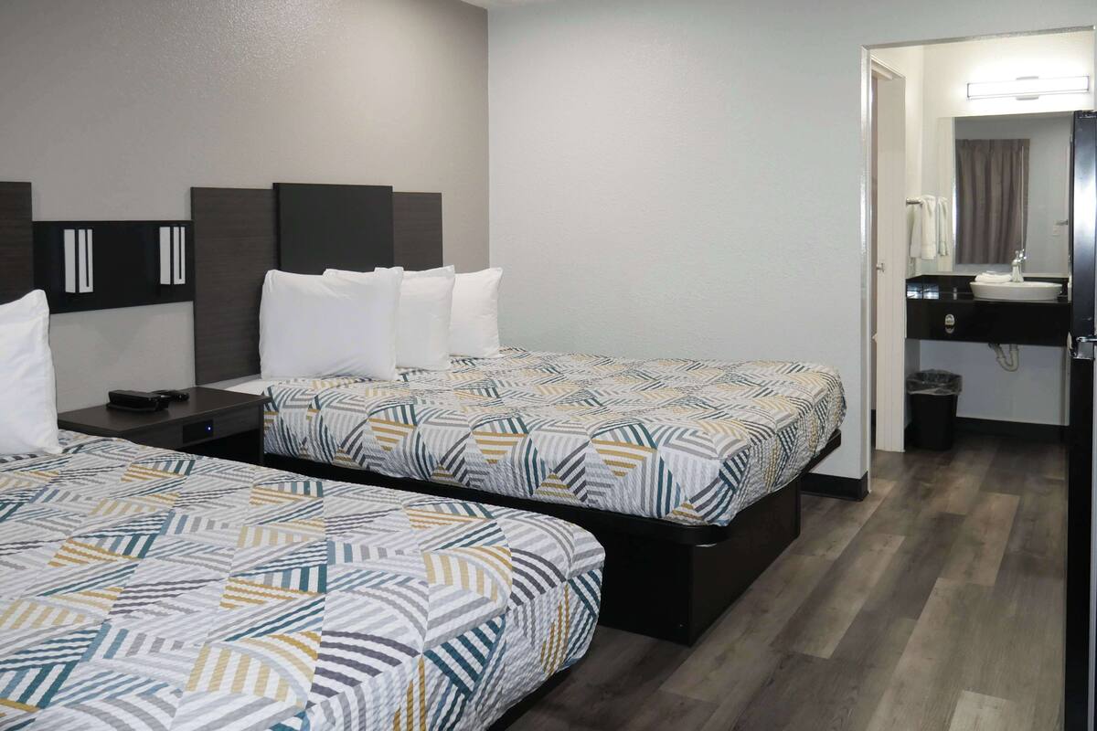 A hotel room with two beds, a bedside table, a mirror, and a bathroom with a sink and mirror.