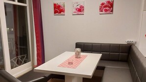 Ein kleiner Essbereich mit einem weißen Tisch, einem Glasbehälter und einer Tischmatte mit Punkten. Zwei schwarze gepolsterte Bänke und ein Fenster mit Vorhang sind ebenfalls vorhanden.