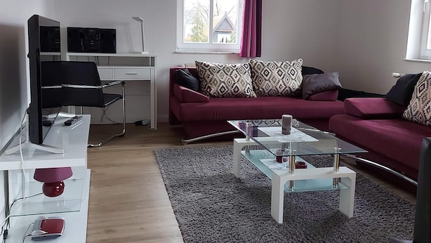 Ein modernes Wohnzimmer mit einem roten Sofa, einem Glastisch, einem weißen Schreibtisch und einem Computerarbeitsplatz.