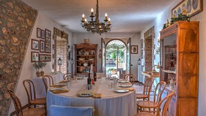 Une salle à manger avec une table ronde dressée pour six personnes, entourée de chaises en osier. Un lustre est suspendu au plafond, et un mur de briques orné de tableaux encadrés se trouve sur le côté gauche.