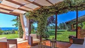 Un espace extérieur aménagé, abrité et doté de mobilier en osier, d’une table avec une bougie et des verres, et offrant une vue sur un jardin luxuriant et des collines lointaines.