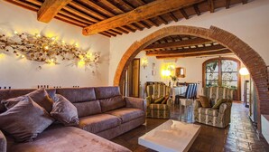 A cozy living room with a brown sofa, patterned armchairs, and a glass coffee table. An arched brick doorway leads to a dining area with a table and chairs. - La Magia 8+2, Emma Villas (Gavorrano)