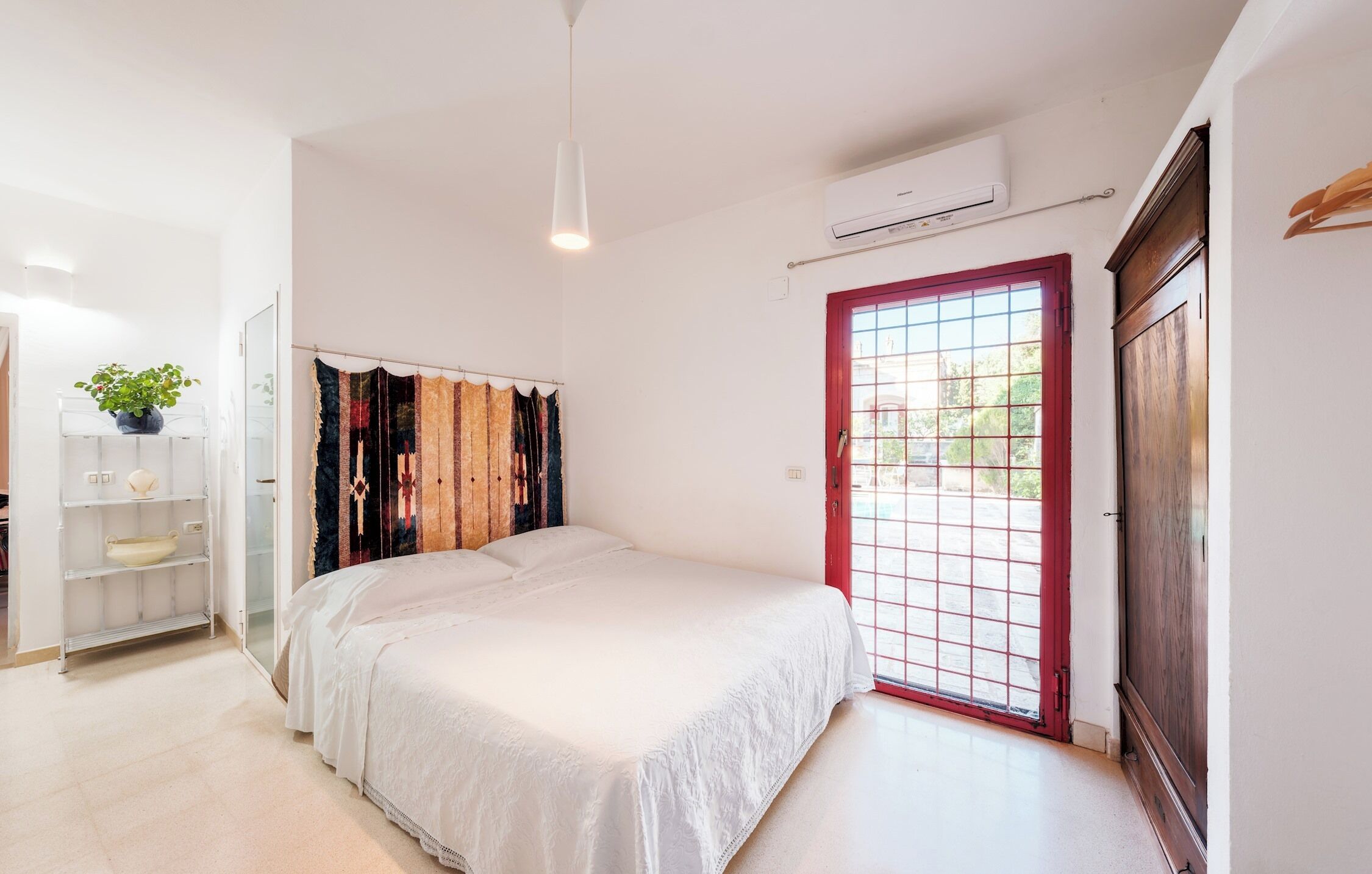 A bedroom with a bed, a wardrobe, a shelf with decorative items, and a window with a red frame.
