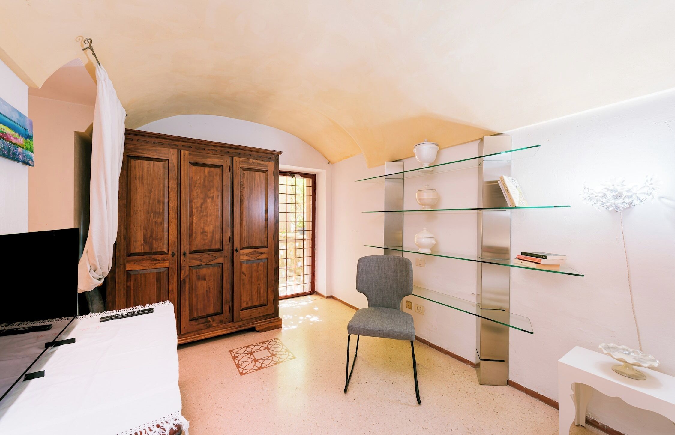A room with a wooden wardrobe, a glass shelf unit, a grey chair, and a white table.