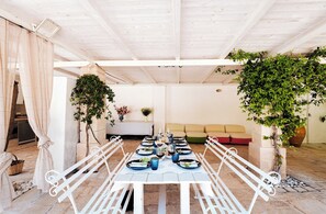 Ein Essbereich mit einem langen Tisch, der für eine Mahlzeit gedeckt ist, umgeben von weißen Metallstühlen. An beiden Seiten des Tisches stehen Kübelpflanzen, und im Hintergrund ist eine Couch zu sehen.