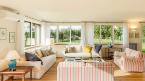 A spacious living room with a large window, a striped ottoman, and a coffee table. - Casale Ombrone 11, Emma Villas (Campagnatico)