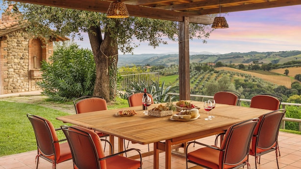 A wooden table with six chairs, a stone building, and a view of rolling hills. - Casale Ombrone 11, Emma Villas (Campagnatico)