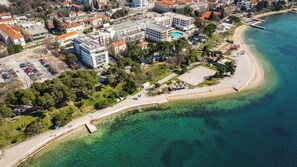 An aerial view of a coastal area with a beach, clear turquoise waters, and a mix of residential buildings and a large hotel with a swimming pool. - Apartment Kolovare (Zadar)