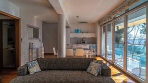 Un salon moderne avec un canapé gris et un coin repas avec des chaises blanches. On aperçoit, au fond, une cuisine avec des meubles blancs et un four à micro-ondes.
