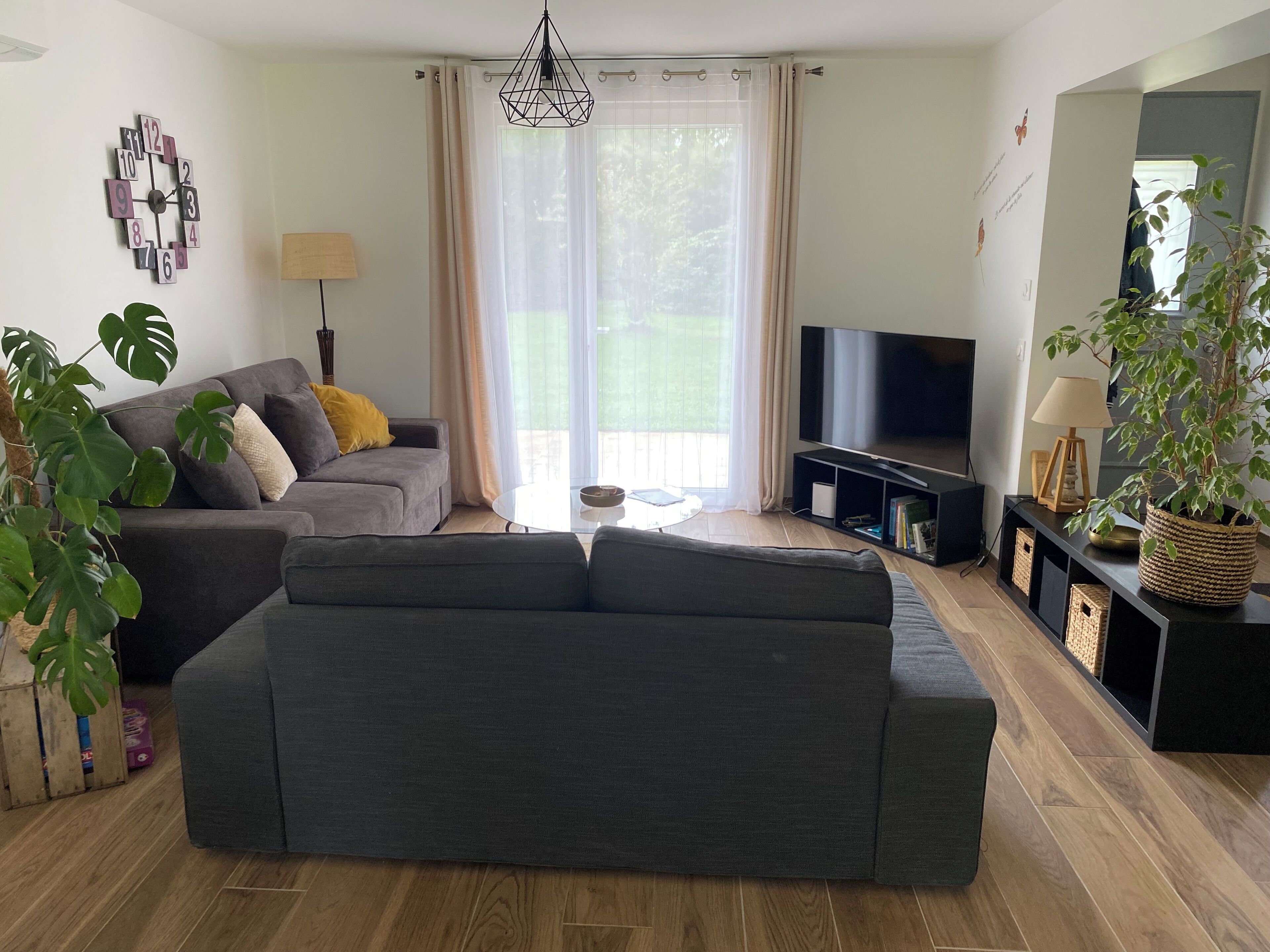 A modern living room with a grey sofa, a flat-screen TV, and a large window with sheer curtains.