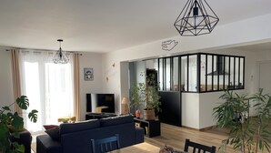 A modern living room with a wooden dining table, dark blue sofa, and a TV. There are potted plants and a geometric light fixture. - Maison l'auvétia 150 m circuit des 24h (Arnage)