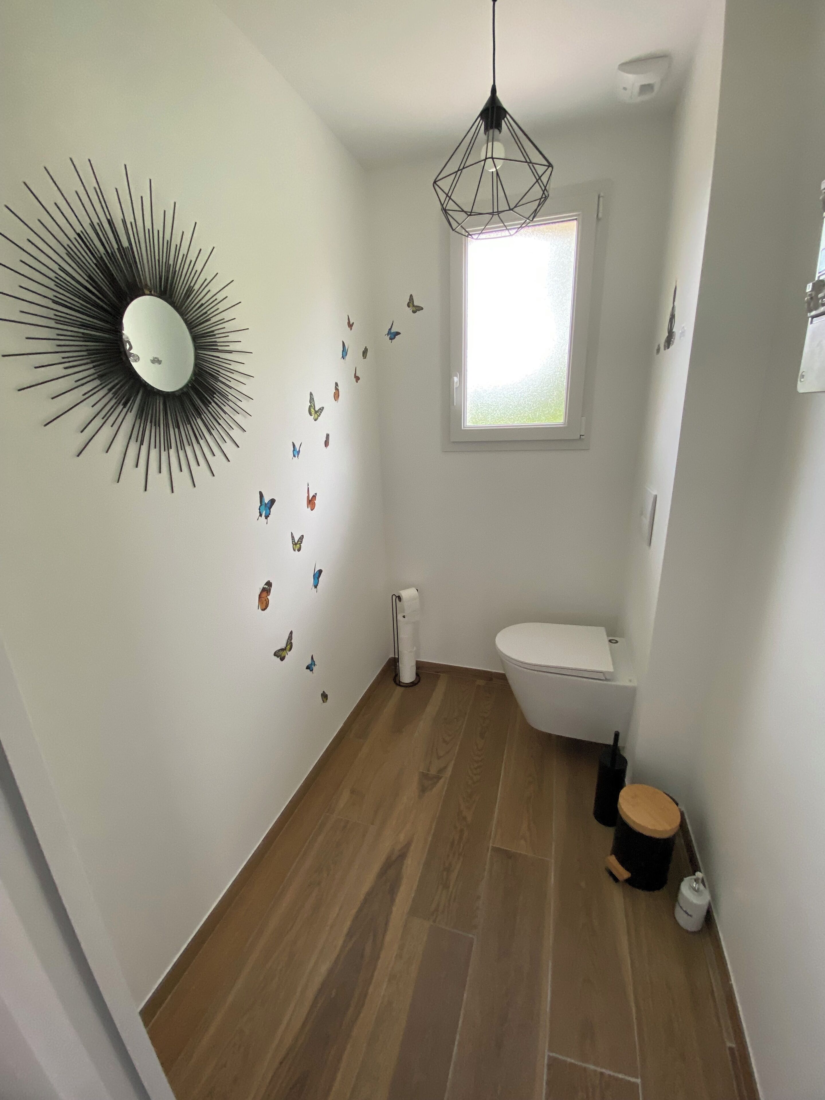 Uma casa de banho com soalho em madeira, sanita branca, candeeiro de teto preto, uma janela e espelho fixado na parede com um design em forma de sol.