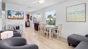 Una sala de estar moderna con comedor, cocina y un amplio ventanal con vistas a palmeras y la playa.