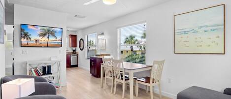A modern living room with a dining area, a kitchen, and a large window with a view of palm trees and the beach.