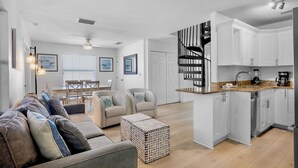 Un salon moderne avec un canapé, des fauteuils et une table basse. On y trouve également un coin cuisine avec des meubles blancs et un escalier en colimaçon noir.