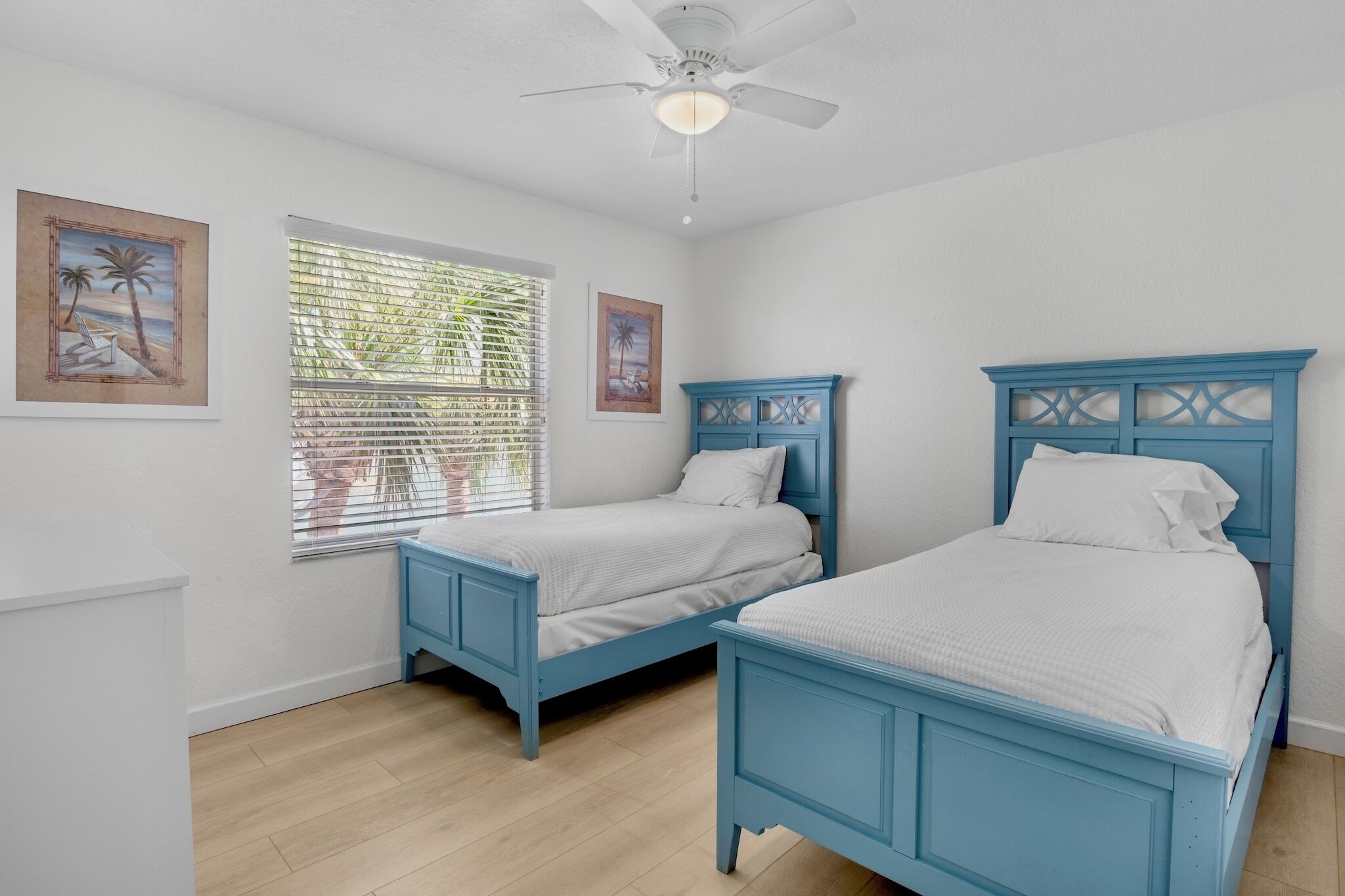 A bedroom with two blue beds, white bedding, a ceiling fan, and two framed pictures on the wall.