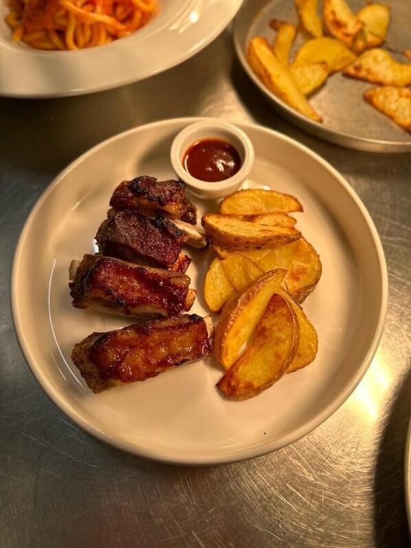 A plate with ribs and potato wedges, accompanied by a small bowl of sauce.