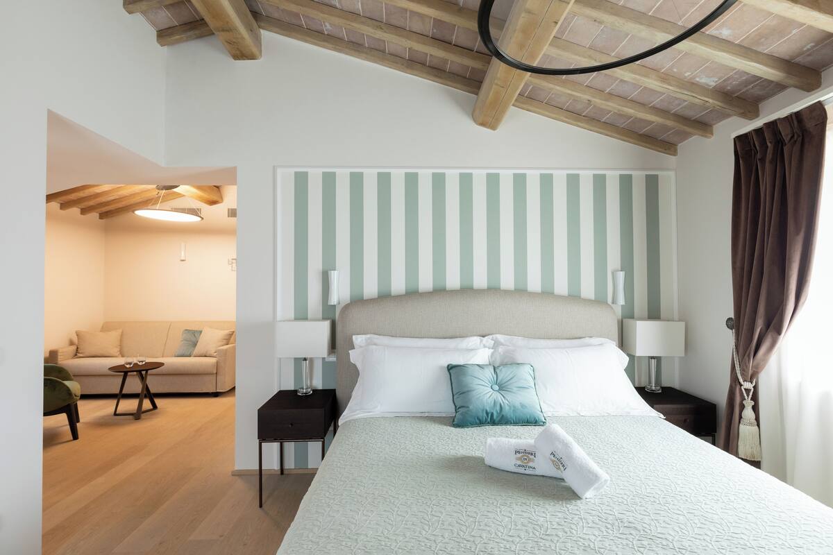 Une chambre d’hôtel avec un grand lit, une tête de lit à rayures, deux tables de chevet avec des lampes, une petite table d’appoint et une vue sur un coin salon avec un canapé et une lampe.