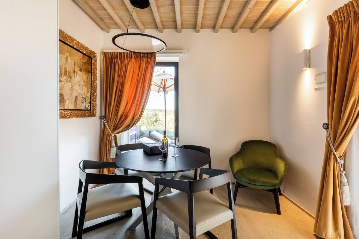 Un coin repas avec une table ronde, des chaises noires, un fauteuil vert et une fenêtre avec des rideaux.