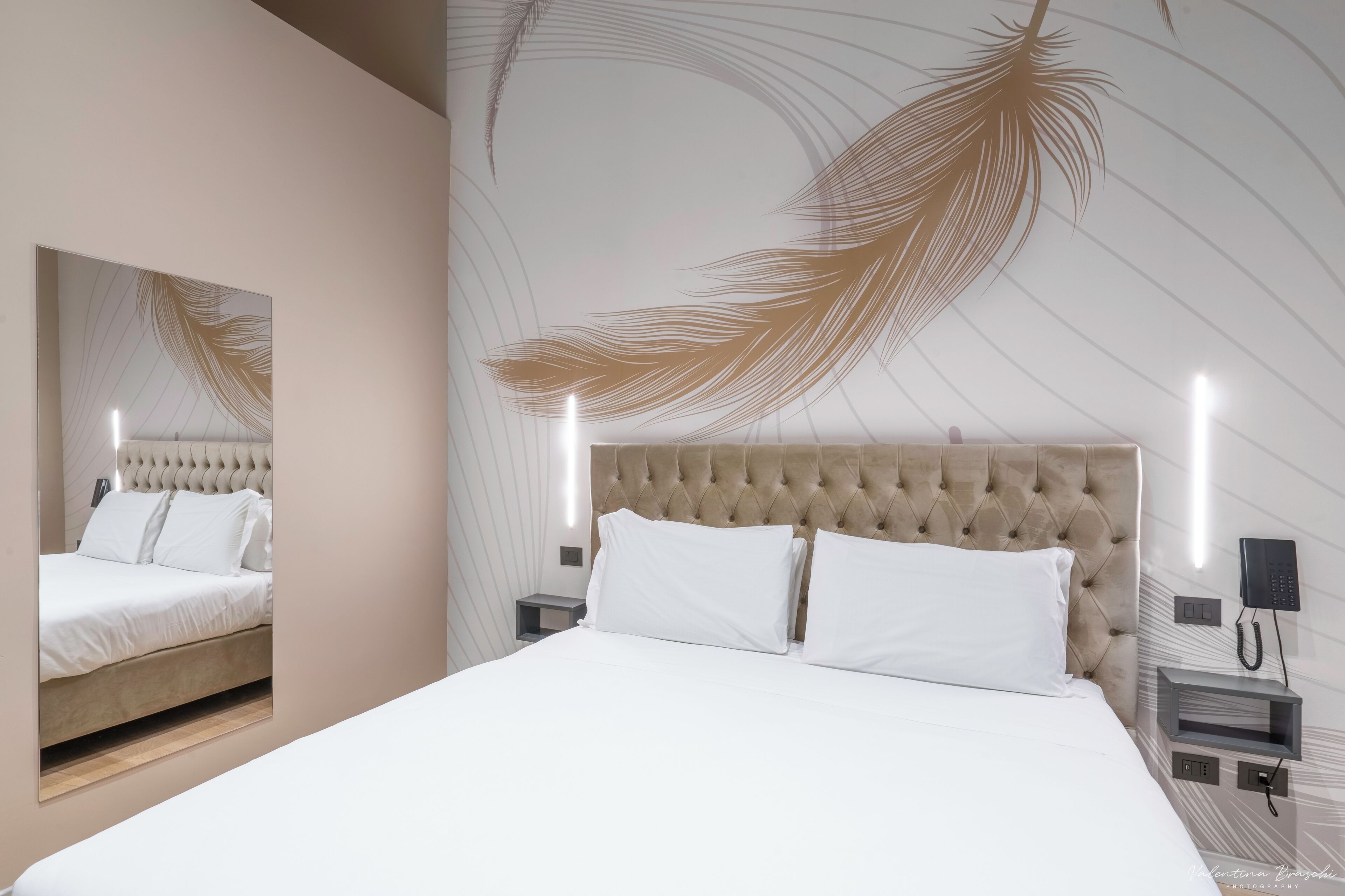 A modern hotel room with a tufted headboard, a large bed with white linens, a nightstand with a phone, and a wall with a feather design.