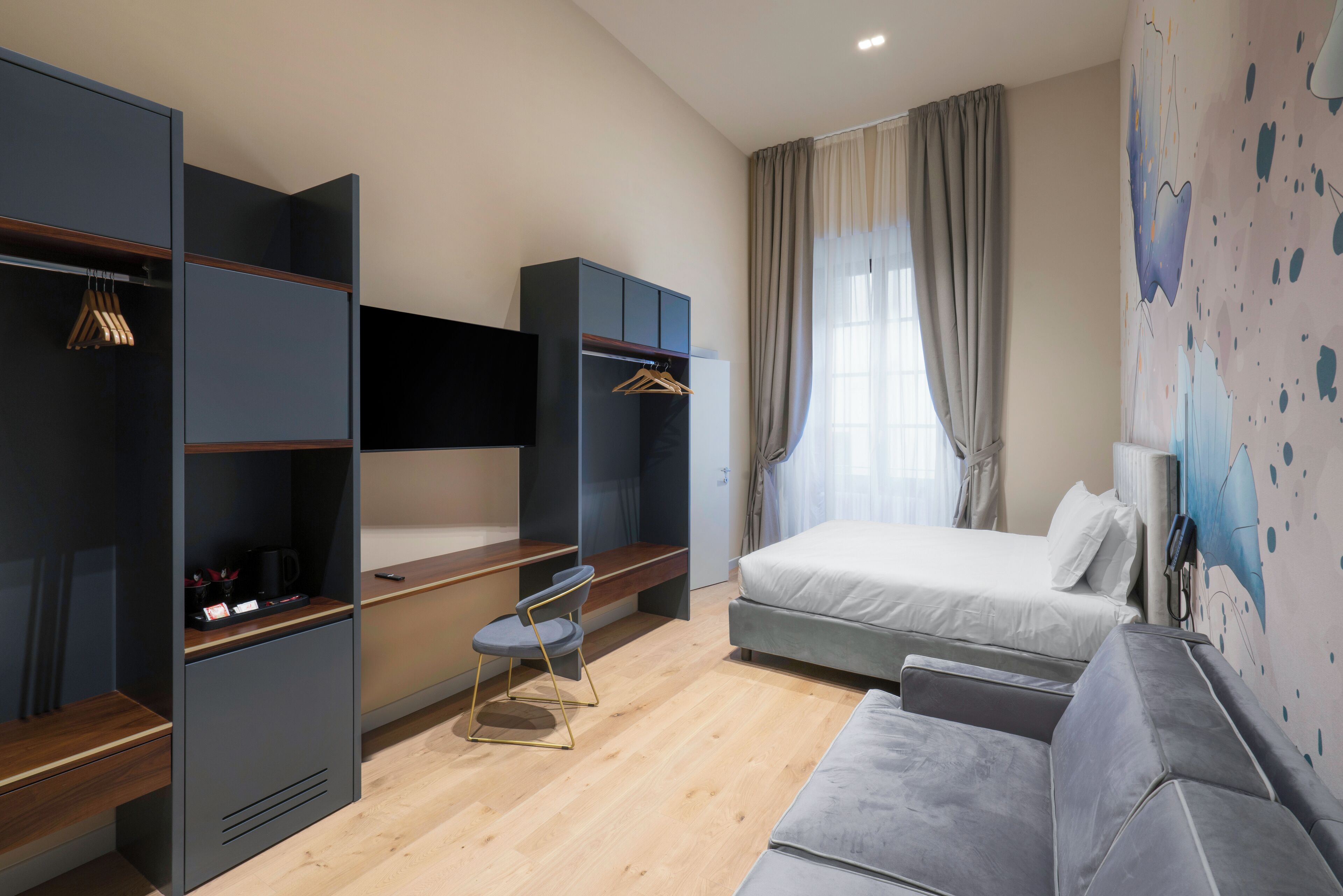 A modern hotel room with a bed, grey sofa, wooden desk, and built-in wardrobe.