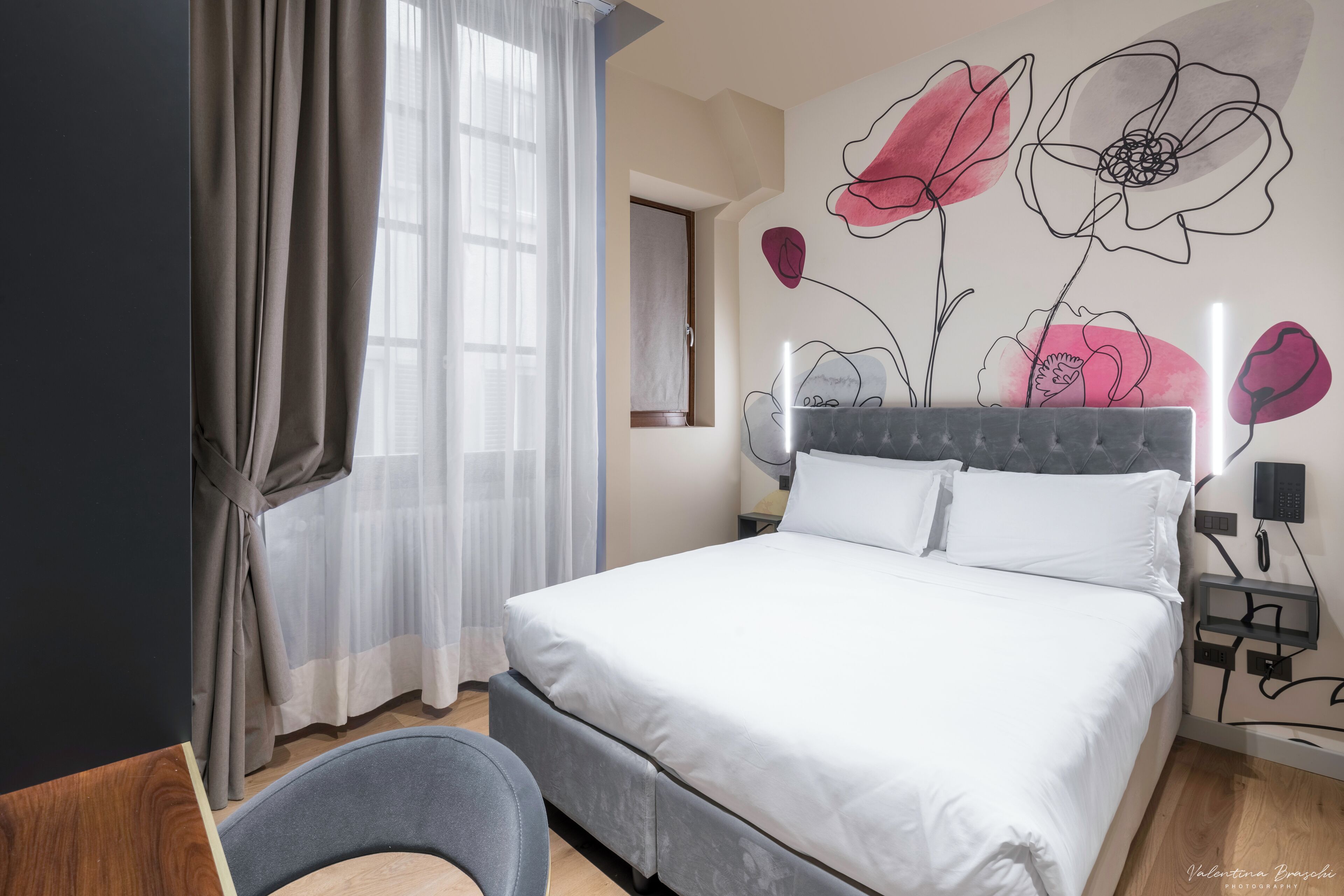 A bedroom with a large bed, a chair, a wall with floral artwork, and a window with curtains.