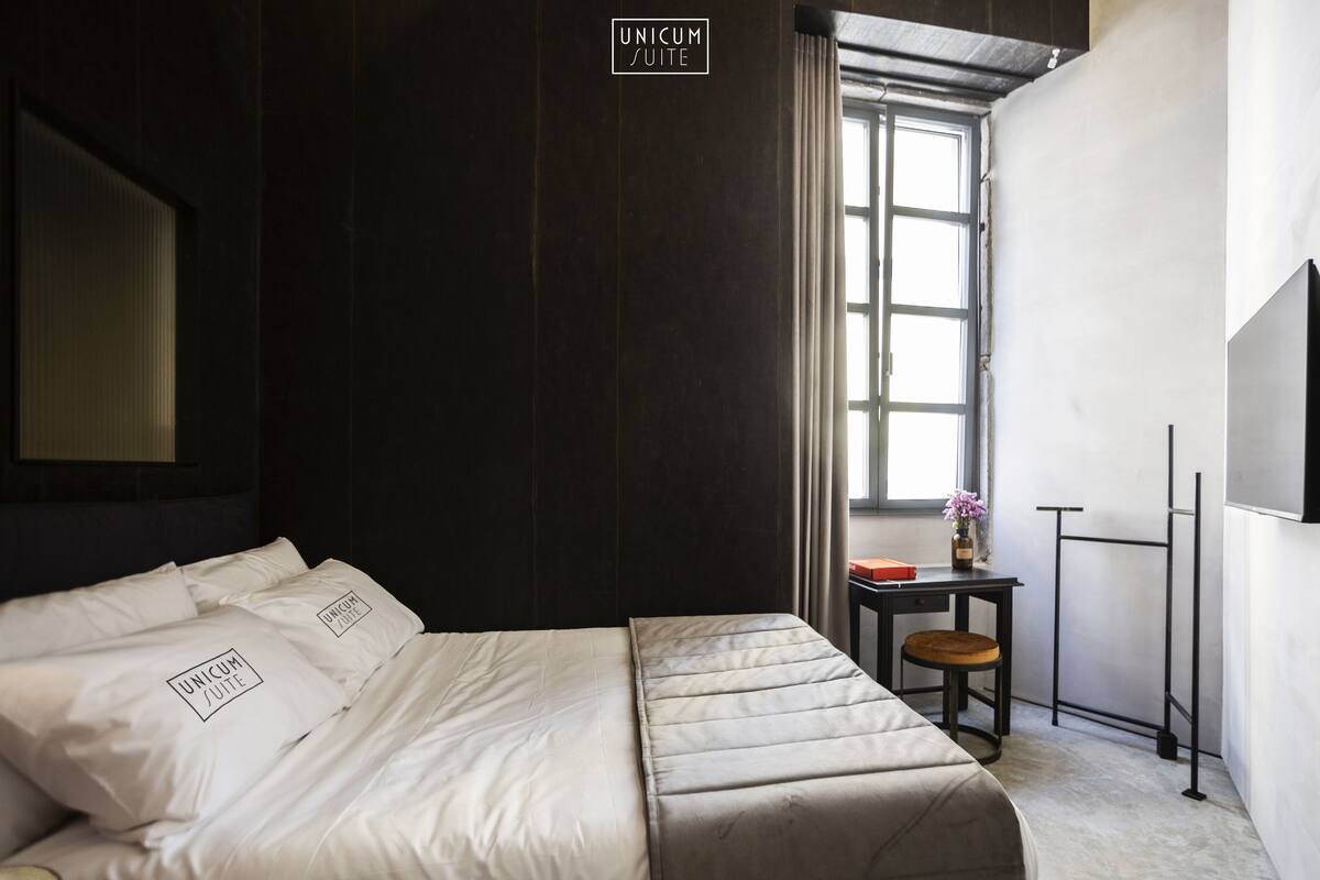 A modern hotel room with a large bed, a small desk, and a window providing natural light.