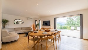 A modern living room with a wooden dining table, beige sofa, and a large window offering a view of the outdoors.