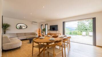 A modern living room with a wooden dining table, beige sofa, and a large window offering a view of the outdoors.