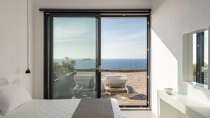 A bedroom with a bed, a glass door leading to an outdoor seating area with a view of the sea.