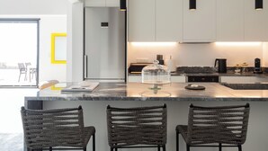 A modern kitchen with a marble countertop, three wicker chairs, and pendant lighting.