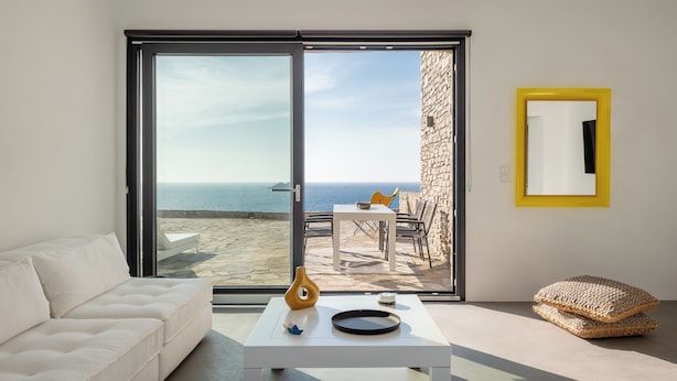 A modern living room with a white sofa, a coffee table with decorative items, a yellow-framed mirror, and a view of the sea through large glass doors.